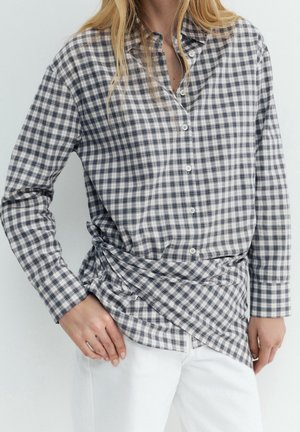 Femme portant une chemise à manches longues à carreaux noirs et blancs avec un détail noué sur un pantalon blanc, posant les mains près des hanches.