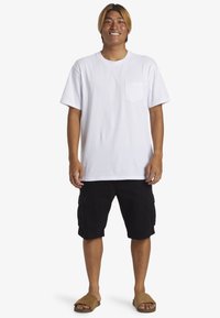 Witte katoenen t-shirt met een borstzak, gecombineerd met zwarte cargoshorts. Schoeisel: beige instapsandalen. Model staat in een neutrale pose.