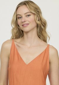 Robe sans manches de couleur rouille avec un décolleté en triangle, fabriquée en tissu texturé. Elle présente des bretelles fines et une coupe décontractée.