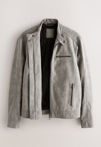 Veste en daim gris clair avec doublure intérieure noire matelassée, poches poitrine et latérales zippées, et détail col à boutons-pression.