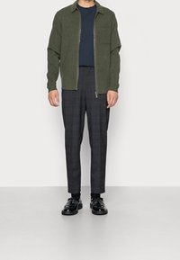 Man med en mörkgrön zip-up jacka, marinblå skjorta, rutiga mörka byxor, svarta strumpor och svarta tassellofers mot en enfärgad bakgrund.