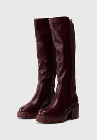 Vīna sarkanas, līdz ceļiem garas ādas zābaki ar biezu papēdi, sānu rāvējslēdzēju un teksturētu zolīti papildu saķerei un stabilitātei.