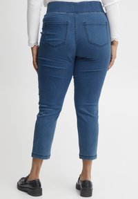 Pantalones de mezclilla en un tono azul medio, con una cintura alta, cinco bolsillos, piernas ajustadas y una sutil textura elástica. Vista trasera.