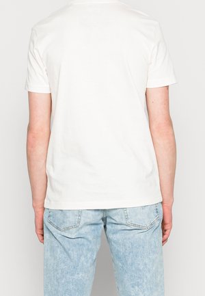 Homme portant un t-shirt blanc uni à manches courtes et un jean en denim bleu clair, vu de dos sur un fond neutre.