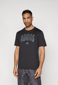 Svart atletisk t-skjorte med "ADIDAS"-logo i grått, rund hals, korte ermer og lett stoff; brukt sammen med mønstrede shorts.