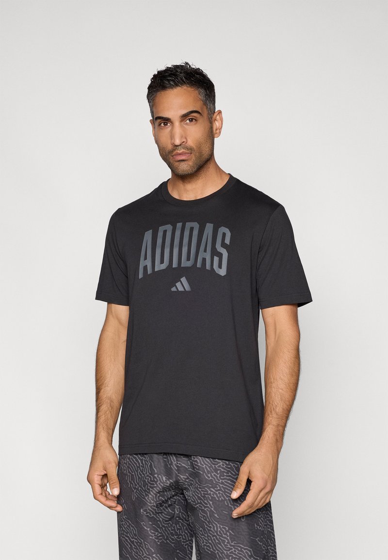 Svart atletisk t-skjorte med "ADIDAS"-logo i grått, rund hals, korte ermer og lett stoff; brukt sammen med mønstrede shorts.