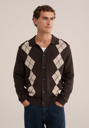 Jeune homme portant un cardigan marron boutonné avec un motif losanges beige sur une chemise blanche, debout les mains dans les poches sur un fond uni.