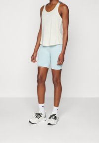 Frau in einem beigen Puma-Tanktop, hellblauen Radshorts, weißen Puma-Socken und weißen Turnschuhen mit schwarzen Schnürsenkeln, die vor einem schlichten Hintergrund steht.