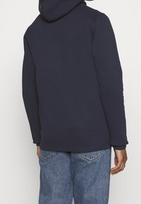 Tmavě modrá mikina s kapucí a hladkým povrchem, se žebrovanými manžetami a lemem. Kombinováno s džíny světle modré barvy, které ukazují zadní detaily.