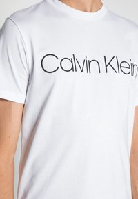 Person wearing a white short-sleeve t-shirt with "Calvin Klein" printed in large black letters across the chest.