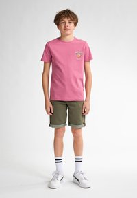 Camiseta de algodón rosa con un gráfico de "Key West" en el pecho; pantalones cortos verde oliva con puños doblados; calcetines blancos con rayas negras; zapatillas blancas.