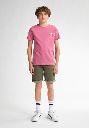 T-shirt in cotone rosa con grafica "Key West" sul petto; pantaloni shorts verde oliva con risvolti; calze bianche con righe nere; sneakers bianche.