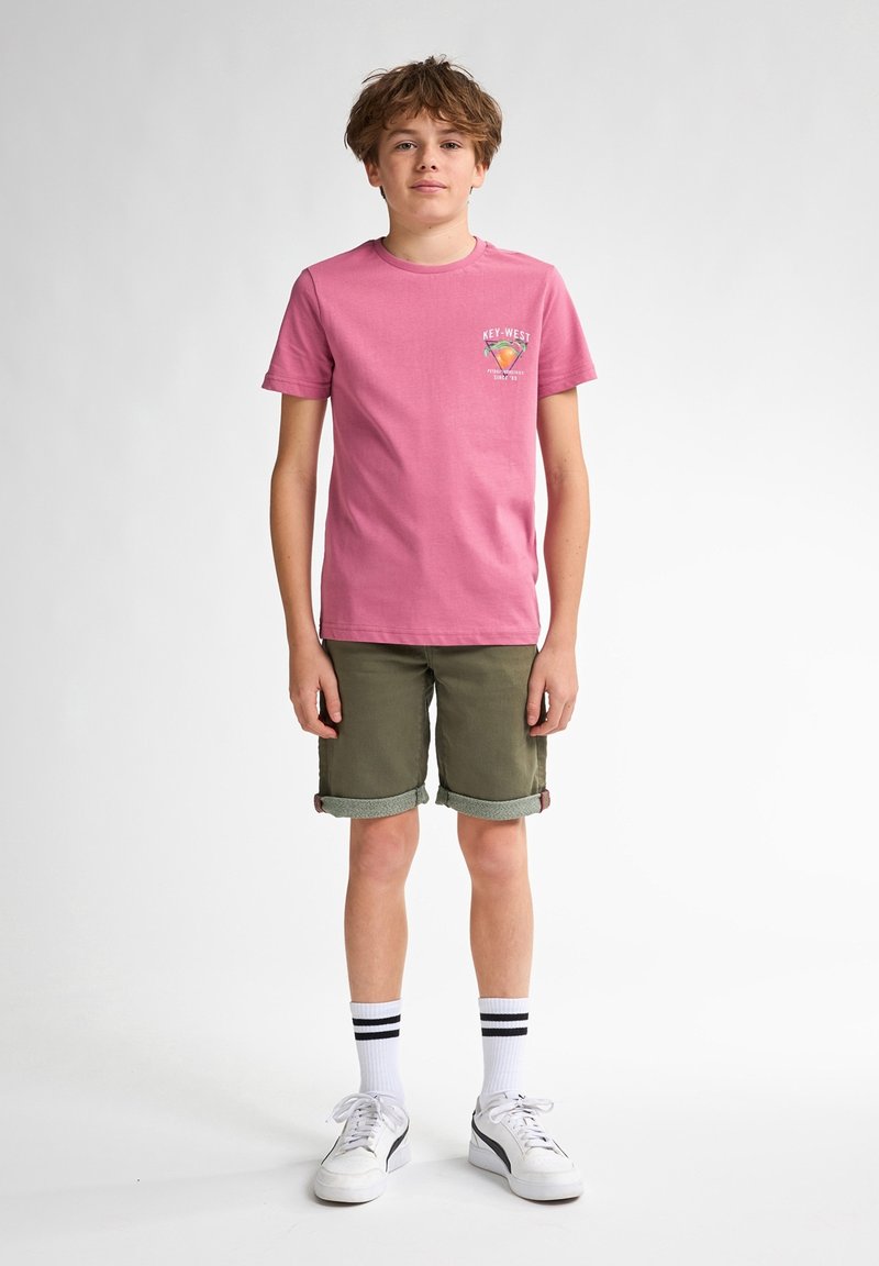 Camiseta de algodón rosa con un gráfico de "Key West" en el pecho; pantalones cortos verde oliva con puños doblados; calcetines blancos con rayas negras; zapatillas blancas.