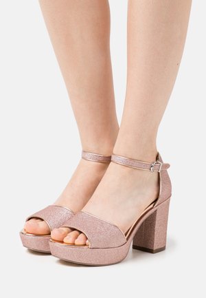 Sandales à talons hauts en paillettes dorées roses avec un design à plateforme, bout ouvert et bride réglable à la cheville. Accents métalliques lisses.