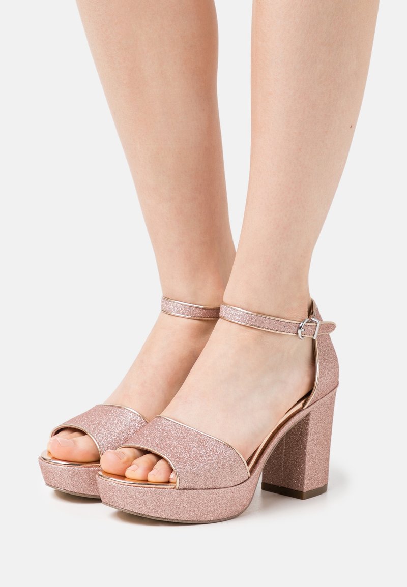 Różowo-złote błyszczące sandały na wysokim obcasie z platformą, otwartym palcem i regulowanym zapięciem na kostkę. Gładkie metaliczne akcenty.