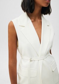 Weißes ärmelloses Blazer mit tiefem V-Ausschnitt, Gürtel-Detail, zwei Vordertaschen und tailliertem Schnitt. Glatter Stoff und strukturierte Gestaltung.