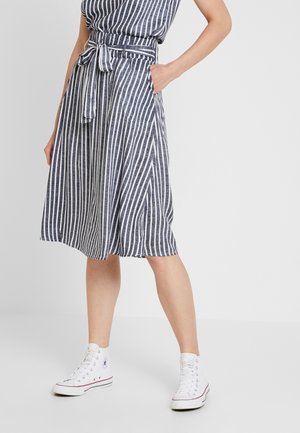 Femme portant une robe rayée verticale bleu marine et blanche longueur genoux, avec ceinture à la taille, mains dans les poches et baskets montantes blanches.