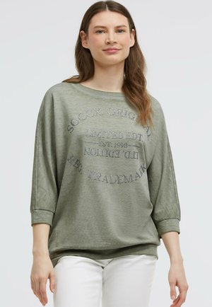 Young woman wearing a loose olive green sweatshirt with silver text and white pants, standing against a plain light background.