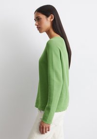 Pull vert à manches longues avec un col rond, texture côtelée et coupe décontractée, associé à un pantalon de couleur claire. Design simple.