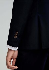 Manga de chaqueta de traje azul marino con tejido texturizado, tres botones marrones y un puño blanco asomando por debajo.