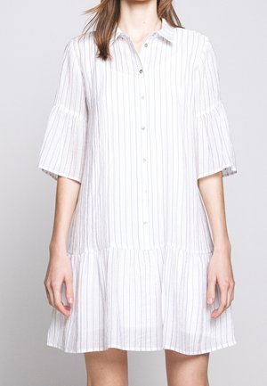 Kobieta ubrana w białą sukienkę zapinaną na guziki, z cienkimi pionowymi niebieskimi paskami i szerokimi rękawami do łokcia, na jednolitym tle.