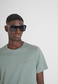 Occhiali da sole con una montatura nera audace e angolare e lenti scure, indossati con una maglietta a maniche corte verde chiaro con un logo discreto.
