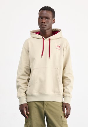 Sudadera beige con capucha de doble capa, cordones rojos y un pequeño logo en el pecho. Tiene un bolsillo frontal y puños y dobladillo acanalados.