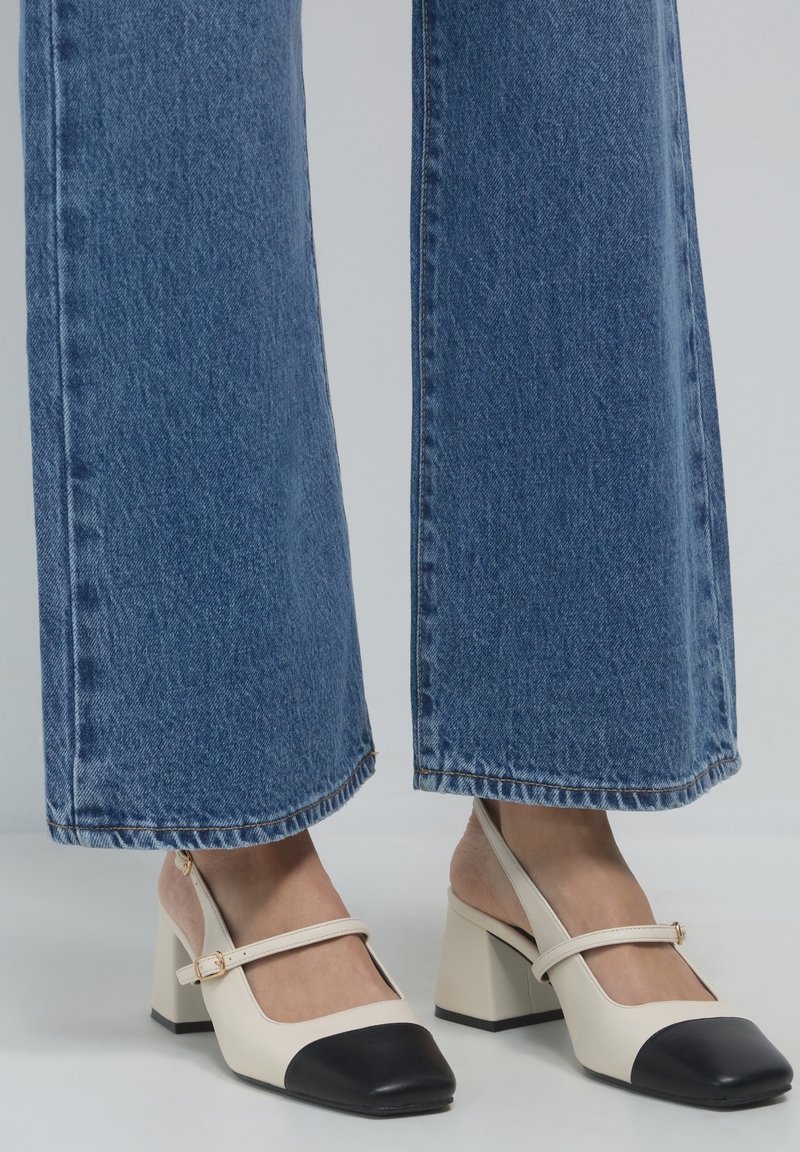 Des chaussures à talons blocs en cuir beige et noir, avec un bout renforcé et une sangle ajustable. Portées avec un pantalon en denim bleu à jambes larges.