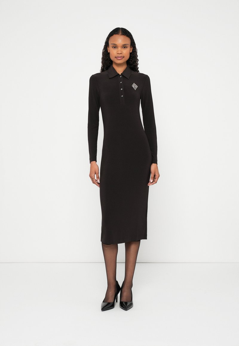 Hosszú ujjú fekete garbóruházat, testhez simuló szabással, gallérral és kis geometriai logóval a mellrészen. Fekete magassarkú cipővel viselve.