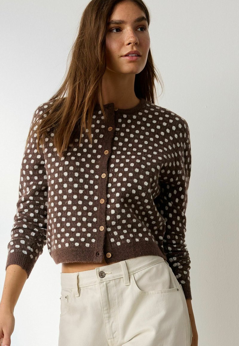 Jeune femme portant un cardigan boutonné à pois marron et un pantalon clair taille haute, debout contre un mur uni.