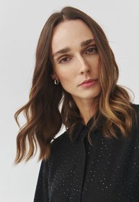 Black, collared shirt with a dotted texture and subtle sheen. Long, wavy hair framing the face. Minimal accessories enhance the look.