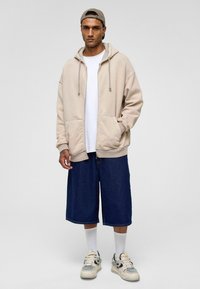 Beige sweatshirt met capuchon en zakken, gedragen over een wit t-shirt. Donkerblauwe denim shorts, witte sokken en grijze sneakers maken de outfit compleet.