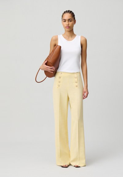 Pantalons large jaunes avec des boutons sur le devant, associés à un débardeur blanc et un sac en cuir marron. Texture lisse et taille ajustée.