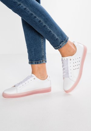 Person iført korte blå jeans og hvide sneakers med pink såler og stjerneformede perforeringer på siden.