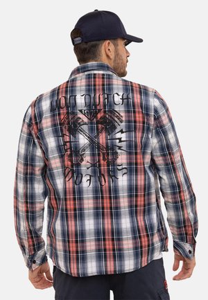 Homme portant une casquette marine et une chemise à carreaux rouge, blanche et bleue avec des pistons croisés et un texte usé au dos, vu de dos.