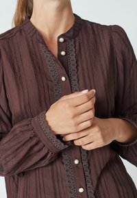 Pruun tekstuuriga pluus vertikaalsete triipudega, ümmarguse kaelusega ja pitsist äärega. Esiküljel on pärlitega nupud ja gathered varrukad.