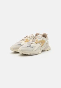 Light beige sneakers with a textured upper, featuring mesh and leather panels, rubber sole, and tonal accents. Lacoste branding on the side.