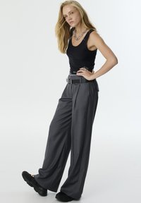 Zwarte tanktop, grijze wijde broek met voorplooien, een zwarte riem met een zilveren gesp en chunky zwarte schoenen. Gladde stoftextuur.