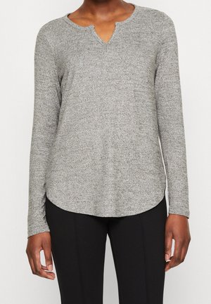 Frau trägt ein graues, langärmeliges Strickoberteil mit V-Ausschnitt und geschwungenem Saum, kombiniert mit schwarzen, eng anliegenden Hosen.