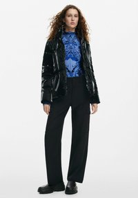 Giacca a vento nera, finitura lucida, silhouette aderente. Sotto, una maglia blu con motivo e collo alto. Pantaloni cargo neri, scarpe con suola spessa.