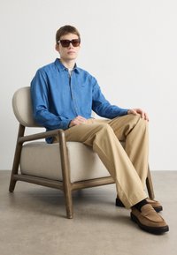 Camicia in denim blu, pantaloni beige, scarpe slip-on marroni, sedia in tessuto grigia, occhiali da sole metallici, sfondo minimalista.