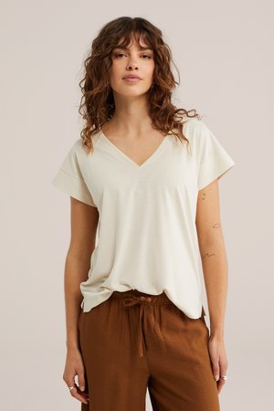 Femme aux cheveux bouclés portant un haut crème à col en V à manches courtes et un pantalon marron avec cordon, sur un fond uni.