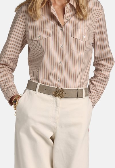 Frau trägt ein beiges und weißes vertikal gestreiftes Hemd, das in eine hoch taillierte cremefarbene Hose mit taupefarbenem Gürtel und goldener Schnalle gesteckt ist.