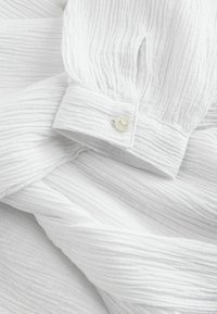 Blouse blanche texturée avec un motif froissé, des manches froncées et une fermeture à boutons avec une finition lisse et nacrée.