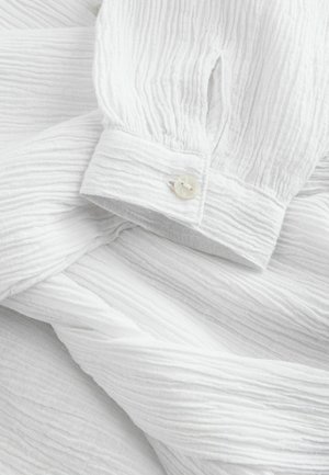 Biała bluzka o fakturowanej powierzchni, z pogniecionym wzorem, marszczonymi rękawami i zapięciem na guziki o gładkim, perłowym wykończeniu.