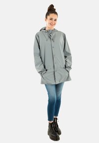 Giacca impermeabile grigio chiaro con chiusura a bottoni, cappuccio e tasche laterali, abbinata a jeans blu e stivali neri con lacci.