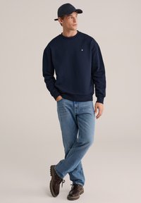 Temno modra majica z okroglim izrezom in nazobčanimi manšetami, kombinirana s svetlo modrimi kavbojkami in rjavimi čevlji, z diskretno podrobnostjo logotipa.