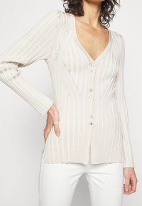 Beige geribde cardigan met een V-hals, sluiting met zes knopen en lange mouwen. Heeft verticale strepen en delicate gaatjespatronen in de stof.
