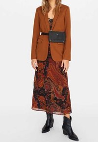 Hnědý blazer se strukturovanými klopami přes černo-oranžové šatové šaty s paisley vzorem, doplněný černými kotníkovými botami a černou páskovou taškou.