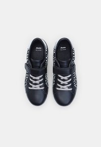 Melni ādas apavi ar baltām punktiņiem, aprīkoti ar Velcro siksniņu, pelēkām šņorēm un teksturētu iekšējo zoli; sportisks dizains ar noapaļotu purngalu.